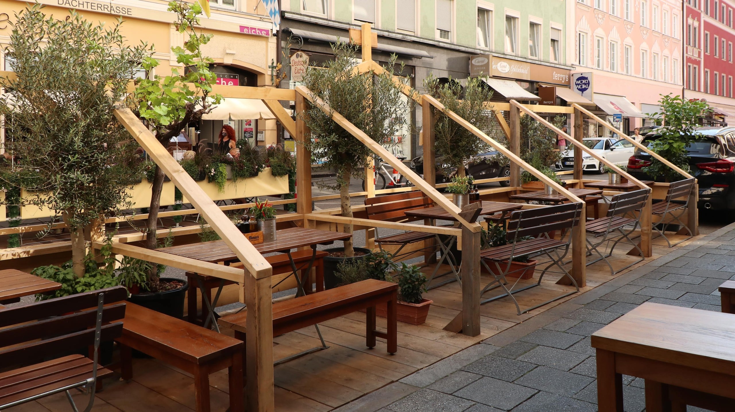 Blick auf einen Schanigarten aus Holz mit mehreren Tischen und Bänken, der in einer Straße in einer Stadt steht, ein Beispiel für temporäres Stadtmobiliar. In vielen Münchner Straßen finden sich inzwischen Schanigärten. Foto: Burkhard Mücke, CC BY-SA 4.0, via Wikimedia Commons
