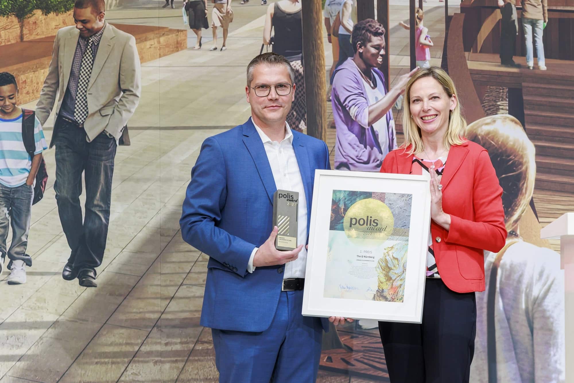 Zwei Personen halten gemeinsam einen Bilderrahmen mit einer Auszeichnung. In der Kategorie „Urbanes Flächenrecycling“ gewann das Projekt The Q in Nürnberg den 1. Preis. Foto: © polis Convention GmbH // Sascha Kreklau