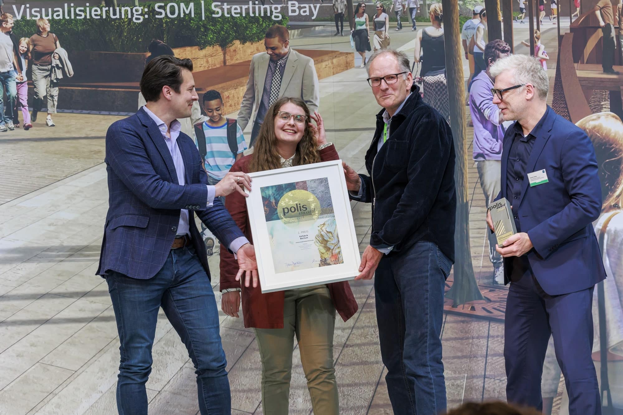 Drei Personen halten einen Bilderrahmen, eine vierte Person steht rechts daneben. Der KoFabrik aus Bochum wurde der 1. Preis in der Kategorie „Soziale Quartiersentwicklung“ verliehen. Foto: © polis Convention GmbH // Sascha Kreklau