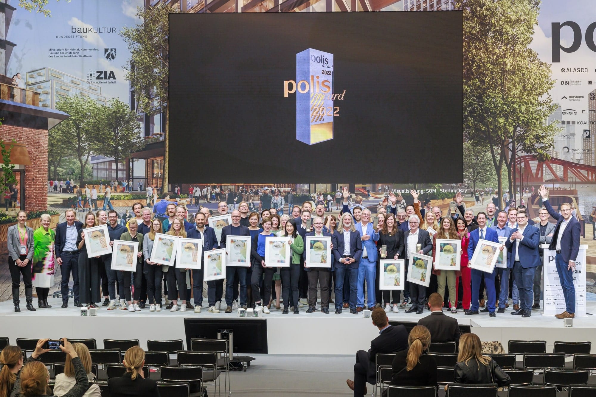 Eine große Gruppe an Personen vor einem großen Bildschirm, auf einer Preisverleihung. polis AWARD 2022, Foto: © polis Convention GmbH // Sascha Kreklau