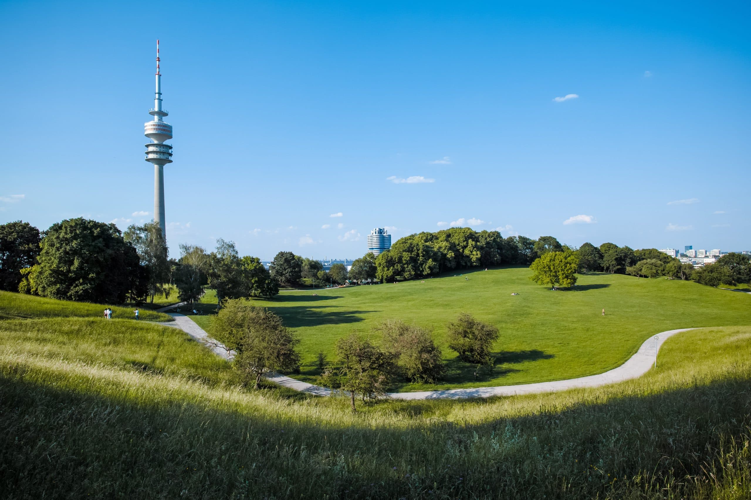 Ansicht von grünen Hügeln mit Bäumen und einem Weg, der sich hoch schlängelt, im Hintergrund der Olympiaturm aus München. Photo: Tuguldur Baatar via Unsplash