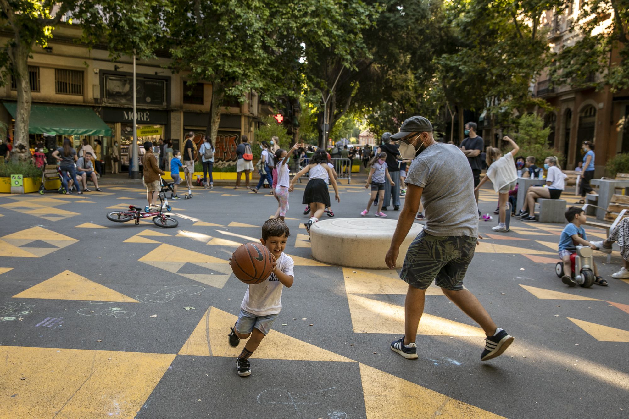 Platz mit schwarz-gelbem Boden mit Dreieck-Muster, spielende Kinder, Erwachsene stehen und sitzen, im Hintergrund Bäume und Gebäude