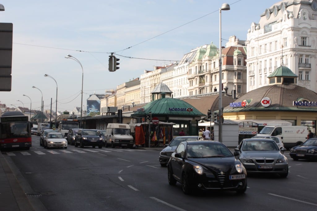 Blick auf den Naschmarkt in Wien, im Vordergrund eine Kreuzung mit mehreren Autos, dahinter die Stände des Markts, im Hintergrund Häuserzeilen. Foto: Politikaner, CC BY-SA 3.0, via Wikimedia Commons