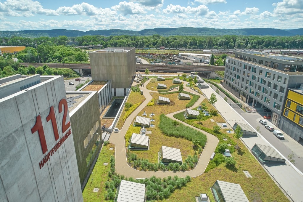 Auf einem Gebäude liegt ein sonnenbeschienener Dachgarten mit einer Wegeführung und unterschiedlichen Pflanzbereichen. Es ist das BuGG Gründach des Jahres 2022 – der genutzte Dachgarten der Hauptfeuerwache Karlsruhe. Credit: Bauder