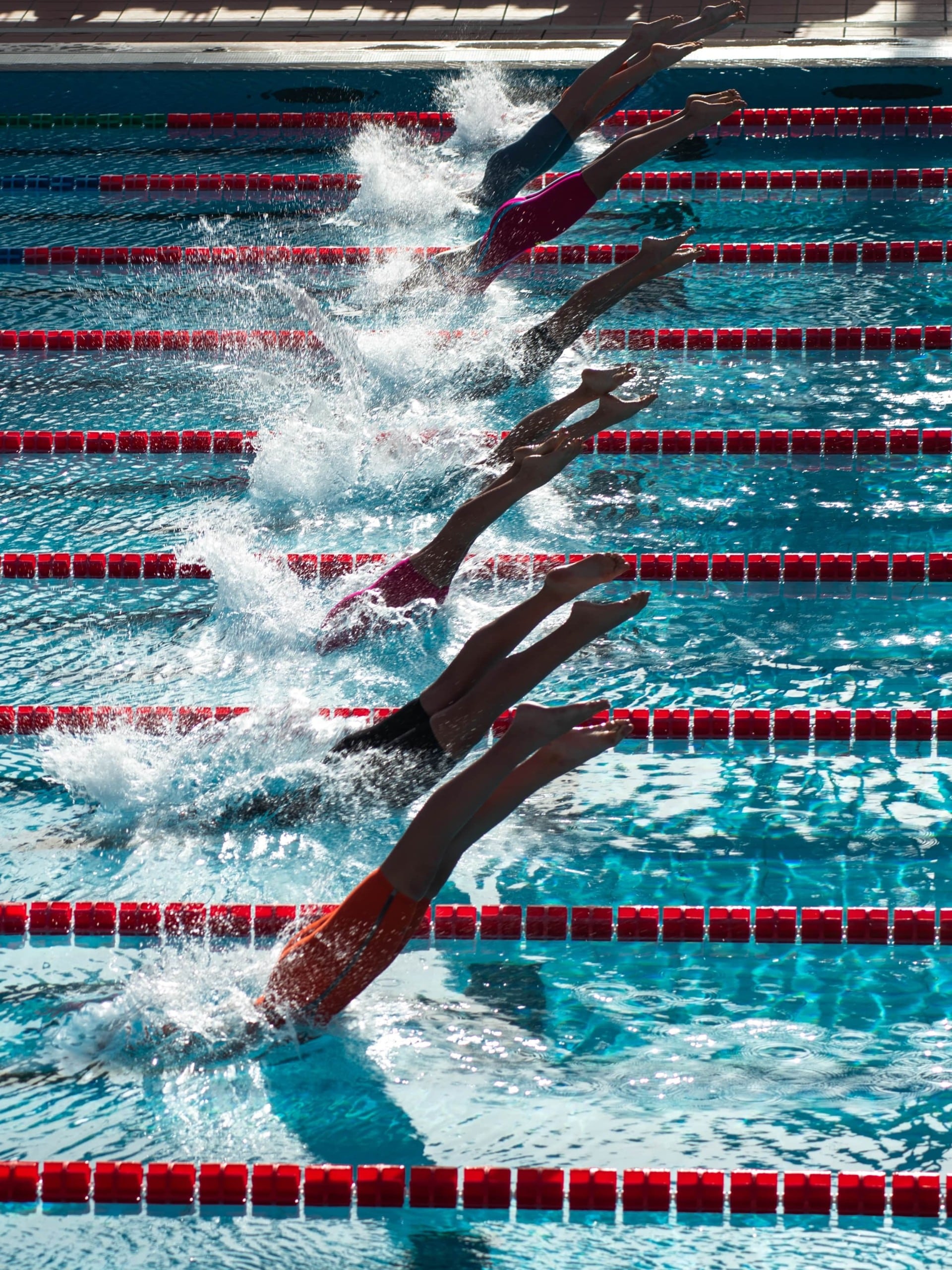 Sieben Personen springen gleichzeitig in nebeneinander liegende Schwimmbahnen eines Pools. Es sind nur noch die Beine zu sehen. Foto: Serena Repice Lentini via Unsplash
