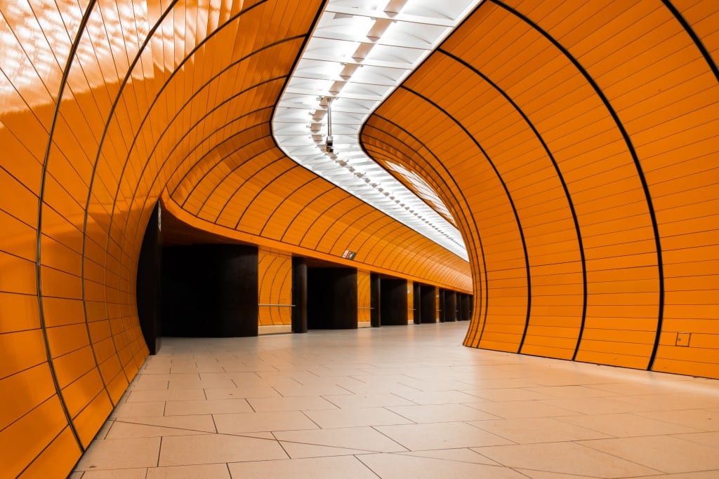 Blick in einen röhrenartigen Gang, dessen Wände mit orangenen Paneelen verkleidet sind, er biegt nach rechts ab. Die Gänge des U-Bahnhofs am Münchner Marienplatz leuchten in markantem Orange. Der S-Bahnhof, an dem mehrere Aktivist*innen die Armlehnen von Sitzbänken abschraubten, liegt darüber. Foto: Nebojsa Pesic via Unsplash