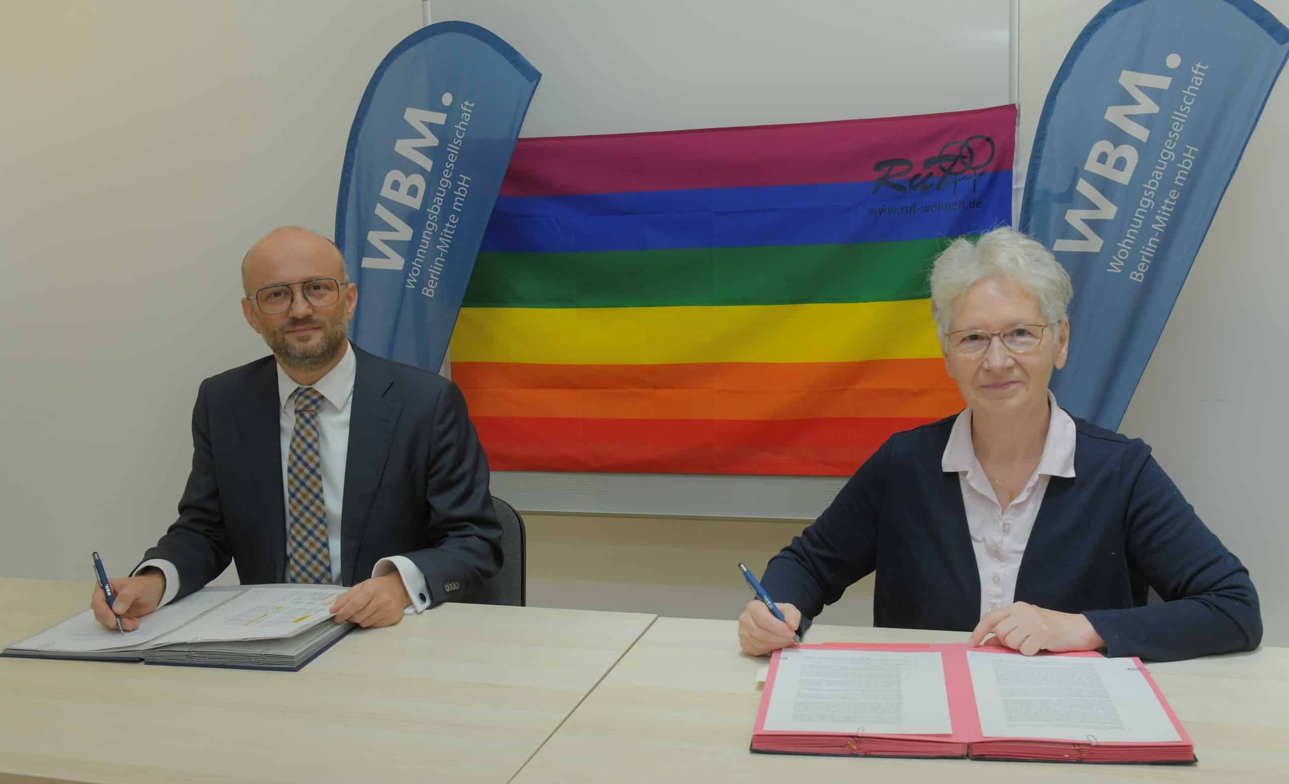 Jutta Brambach, Geschäftsführerin der RuT gGmbH und Steffen Helbig, Geschäftsführer WBM unterzeichnen den Vertrag. Sie sitzen vor einer Regenbogenflagge an einem Tisch. Vor ihnen liegen Vertragsunterlagen.