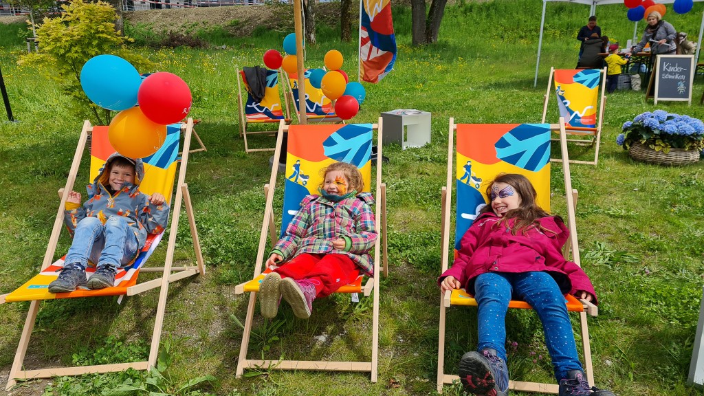 Drei Kinder mit Gesichtsbemalung sitzen in bunten Liegestühlen. Bunte Luftballons im Hintergrund.
