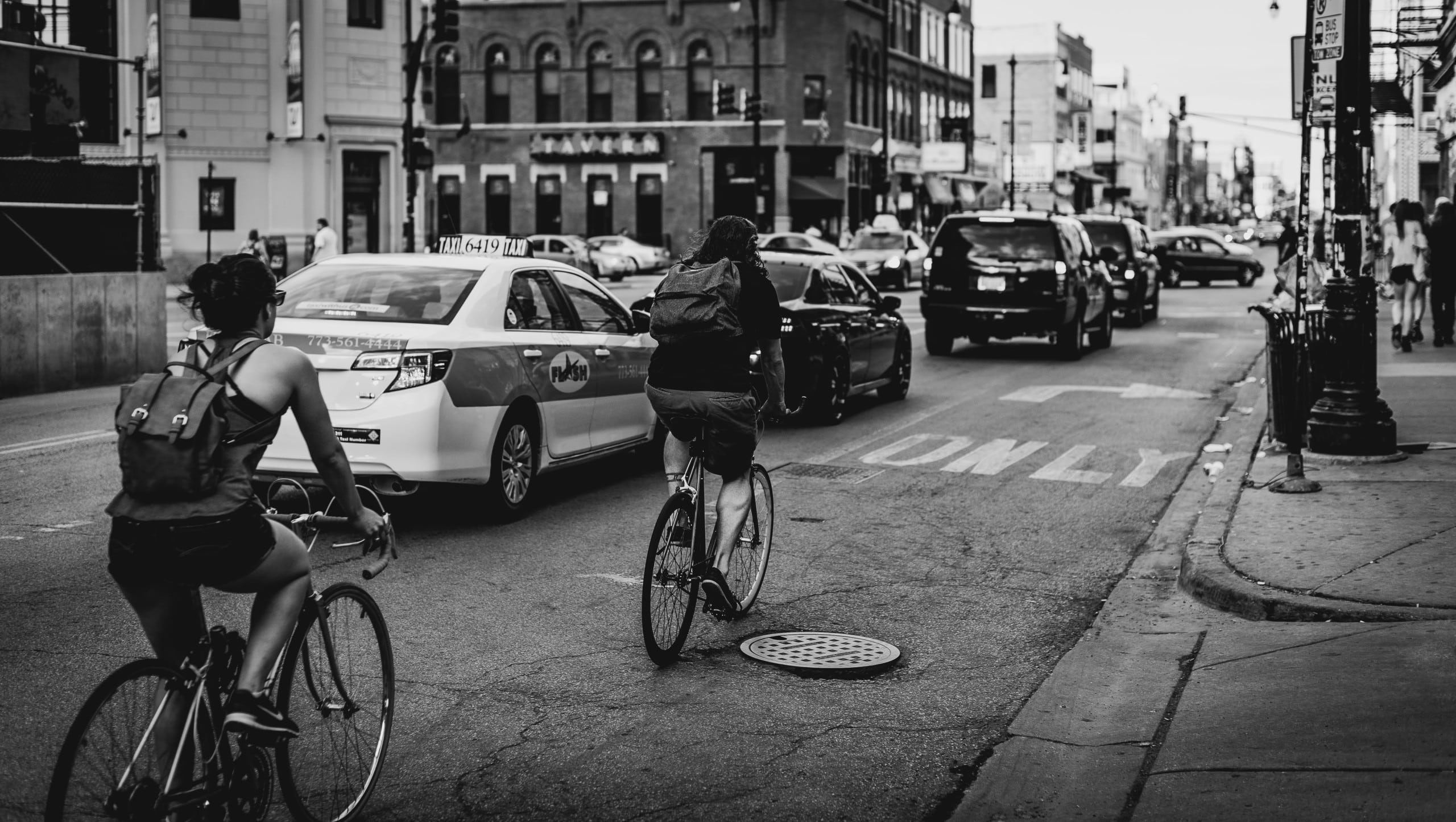 Im Vordergrund zwei Radfahrer, im Hintergrund Autos. In einer städtischen Szenerie.