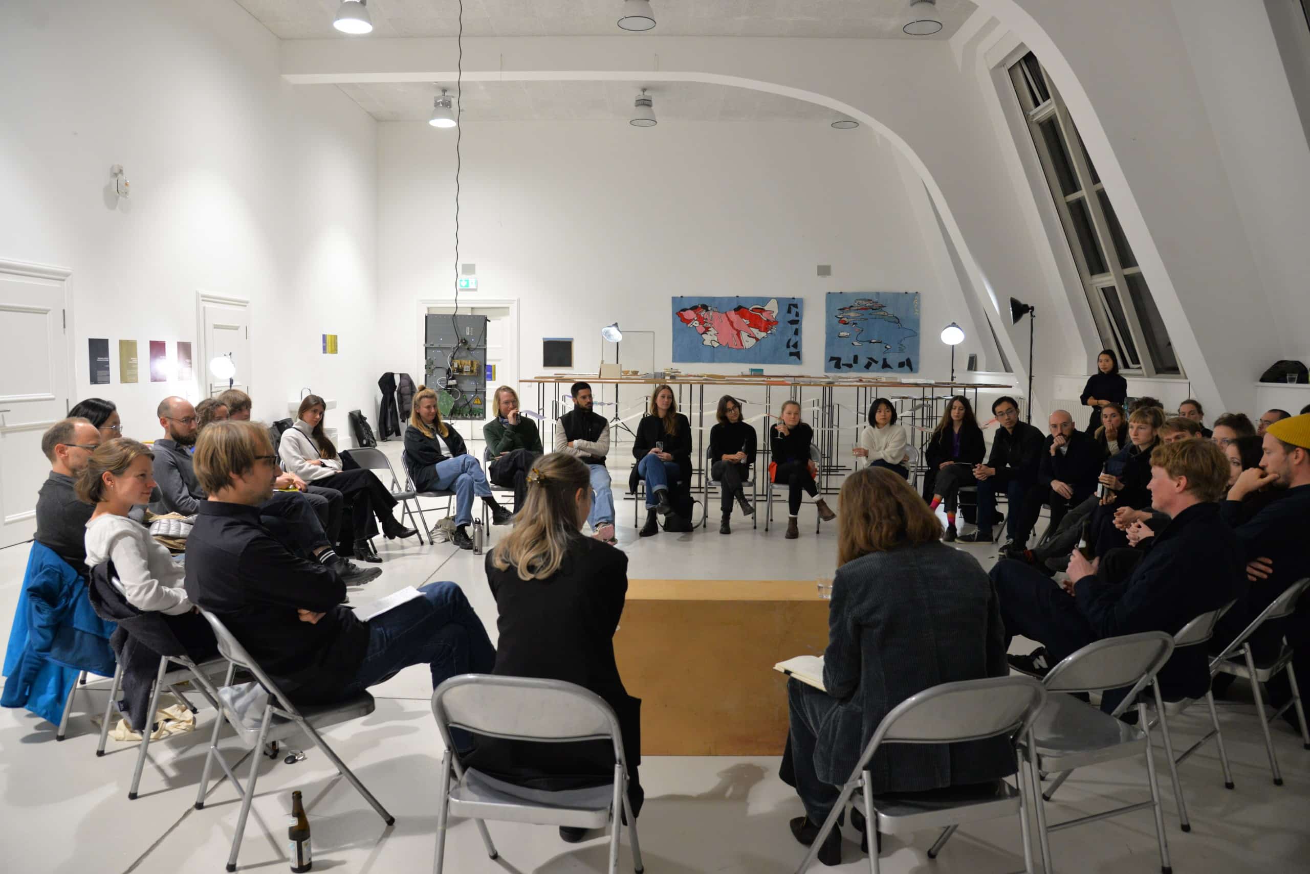 Im Stuhlkreis sitzen diverse Teilnehmer:innen des Symposiums in einem weiß gestrichenen Raum. Im Hintergrund ausgestellte Arbeiten.