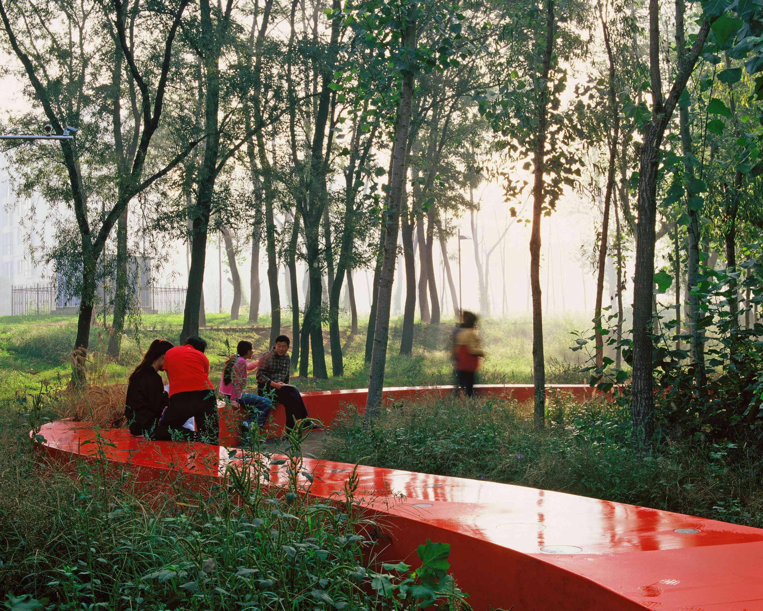Ein rotes Bankelement zieht sich durch einen grünen Park. Menschen sitzen darauf.