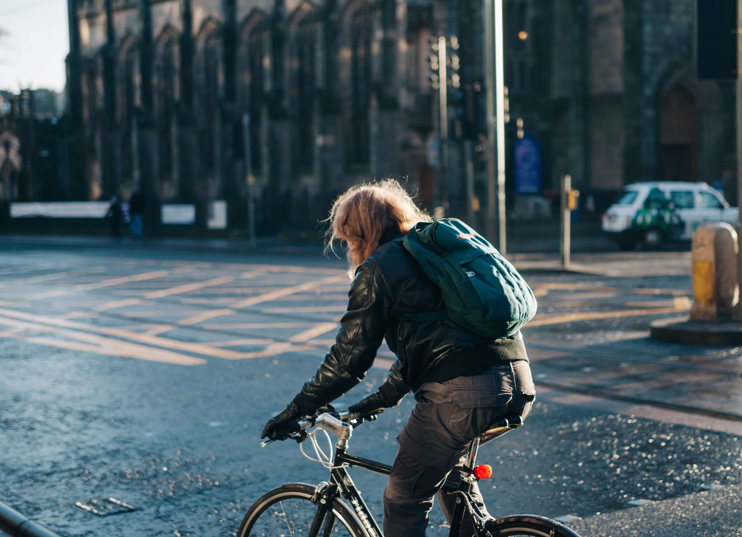Im Vordergrund eine Person mit Rucksack auf einem Fahrrad. Im Hintergrund eine Stadtkulisse mit gepflastertem Belag.